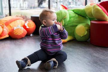 Fototapeta premium Cute fair baby girl sitting in playroom in half-profile on floor with one leg extended sucking on toy pensively, with colourful toys and poufs in the background