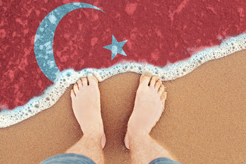 Feet on the sunny sandy beach with flag Turkey. Top View on sea surf.