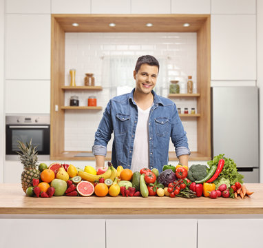 Young Handsome Man With A Variety Of Fruits And Vegetables On A Wooden Counter In A Kitchen