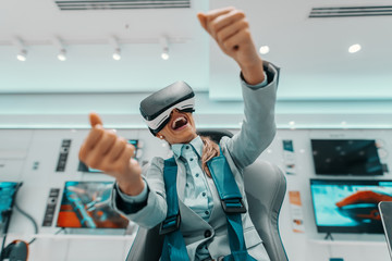 Smiling Caucasian woman in formal wear trying out virtual reality technology while sitting in the chair in tech store.