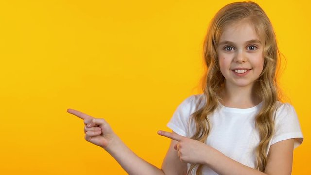 Smiling girl pointing fingers at empty space on yellow background, template