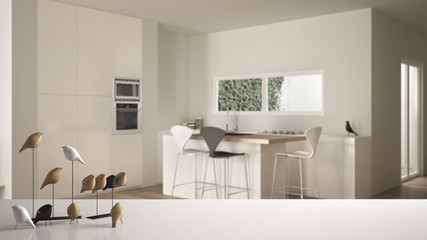 White table top or shelf with minimalistic bird ornament, birdie knick - knack over blurred contemporary white and wooden kitchen with island and stools, modern interior design