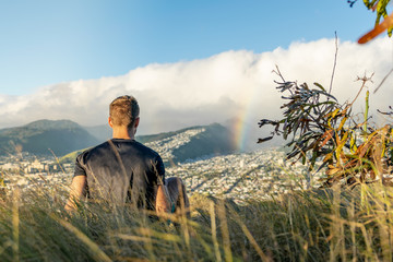 Young male hiker sitting on the summit of Diamond Head Crater in Honolulu on the Island of Oahu, Hawaii shortly before sunset, looking at a rainbow over the city and mountain range behind Honolulu.