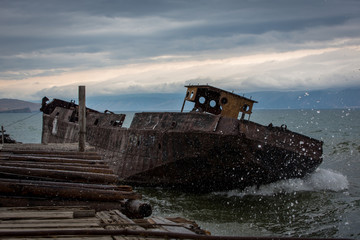 Old, rusty ship near the pier. Big waves flood the deck. Summer cloudy evening.