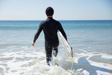 Back view of man carrying surfboard and walking into ocean water