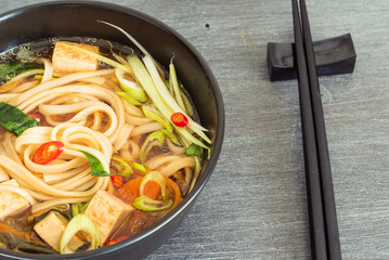 Vegetarian Japanese dietary soup with udon noodles, tofu and vegetables in a black bowl with chopsticks, top view, close-up