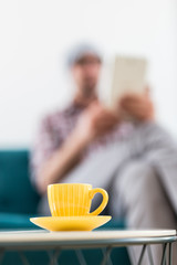 Yellow cup of coffee on small table and fashionable businessman with digital tablet in the background.