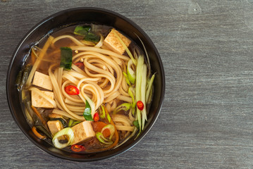 Vegetarian dietary Asian soup with udon noodles, tofu and vegetables in a black bowl, top view, close-up, copy space