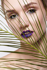 Female portrait with palm branch leaves on foreground and beauty face makeup with violet lips.