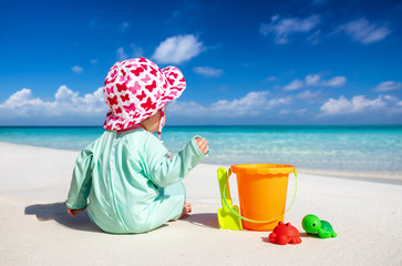 Kleines Baby mit Sonnenschutz Kleidung sitzt am tropischen Strand und hat Spaß mit seinen...