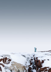a girl in a bright blue jacket with a backpack walks on top of a red rock during a heavy snow storm in the Arctic Circle