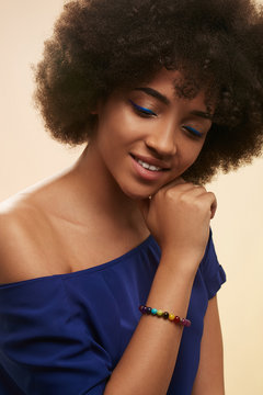 Portrait of African girl with blue eyeliner, wearing T-shirt with nacked shoulder and stony bracelet with multicolored stones. The woman with her head low is touching her neck on beige background.