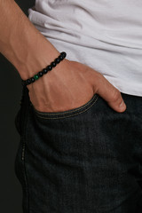 Cropped half-turn shot of guy's hand with stony bracelet with smooth surface on black and malachite beads. The man in jeans and T-shirt is putting his hand into pocket, posing on dark background.