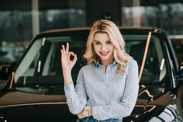 Fototapeta premium cheerful blonde woman showing ok sign near black automobile in car showroom