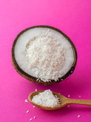 Halves of coconut and small pieces of coconut with woden spoon on a pink background
