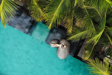 Zelfklevend Fotobehang Top view of slim young woman in beige bikini and straw hat relaxing near luxury swimming pool and palm trees.Vacation concept. Drone photo © tatyanasuyarova