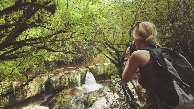 slender girl with blond braided hair stands near dark fence, young tourist with large travel backpack pulls out camera and takes photos of nature. charming beauty of waterfall in Martville canyon