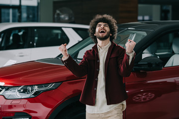 cheerful curly man gesturing while standing near red car