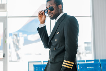 stylish confident african american pilot in sunglasses in departure lounge in airport