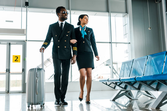 african american pilot and stewardess walking together with suitcase in airport