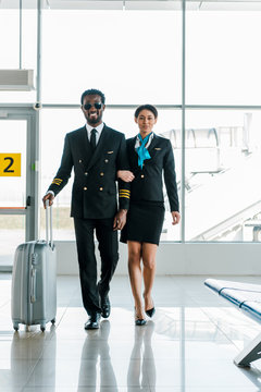 african american pilot and stewardess walking together with baggage in airport