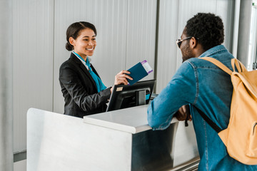 smiling african american airport worker giving passport and air ticket to tourist with backpack