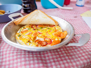 Closeup of fried eggs with sausages and chives in a small steel pan and a piece of buttered toast. Chiang Rai, Thailand. Travel and cuisine.