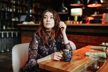 Gorgeous girl with curly hairstyle waiting friend in restaurant with cozy interior and drink cofe.