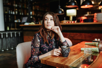 Gorgeous girl with curly hairstyle waiting friend in restaurant with cozy interior and drink cofe.