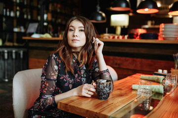 Gorgeous girl with curly hairstyle waiting friend in restaurant with cozy interior and drink cofe.