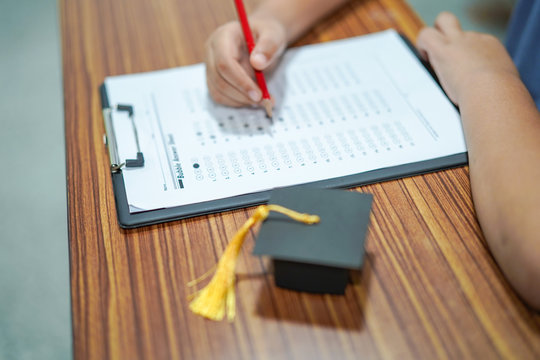Asian children young kid write answer in a quiz examination test study learn with graduation gap hat on wooden table in school : bright genius education concept.