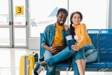 happy african american couple sitting in departure lounge with suitcase and air tickets in airport