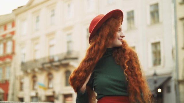 Happy smiling redhead lady with natural freckles, long curly hair posing in street of the city