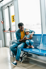 african american father sitting in waiting hall in airport and playing with son