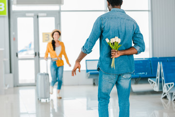 selective focus of african american man walking towards happy girlfriend with suitcase while hiding tulips behind his back in airport