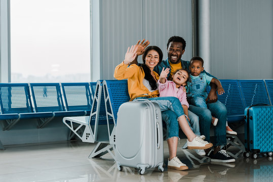 smiling african american family with baggage and kids sitting in airport and waving hands together