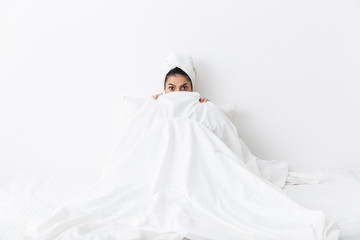 Scared amazing woman with towel on head lies in bed covering herself under blanket isolated over...