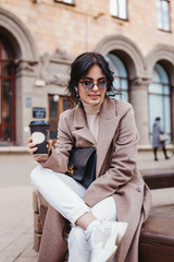 Beautiful young girl in coat and sunglasses sitting on the bench. Woman drinking coffee and laughing.