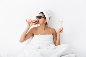 Emotional woman with towel on head lies in bed under blanket isolated over white wall background wearing sunglasses eat burger.