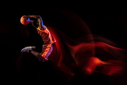Heating the atmosphere. African-american young basketball player of red team in action and neon lights over dark studio background. Concept of sport, movement, energy and dynamic, healthy lifestyle. - Powered by Adobe