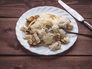 Rice with seafood, squid, mussels with vegetable oil on a white plate. Fork and knife on a wooden plank tray.