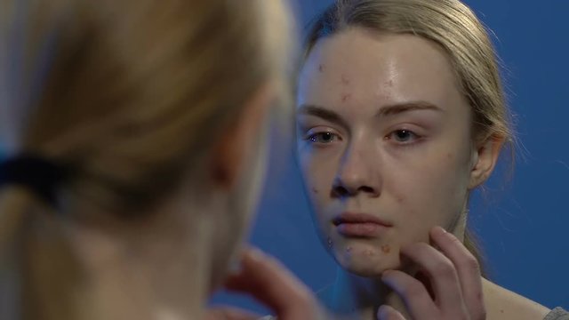 Young crying girl looking at face acne in mirror, suffering from skin problems
