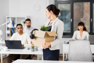 business, firing and job loss concept - sad fired female employee with box of personal stuff leaving office looking at her colleagues