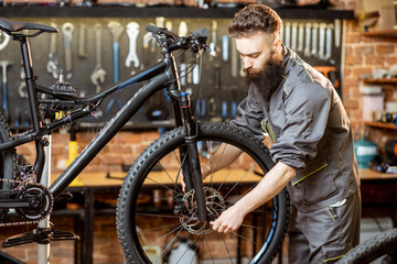 Fototapeta na wymiar Handsome bearded repairman in workwear mounting wheel on a mountain bicycle at the workshop
