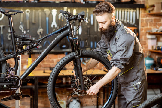 Handsome bearded repairman in workwear mounting wheel on a mountain bicycle at the workshop
