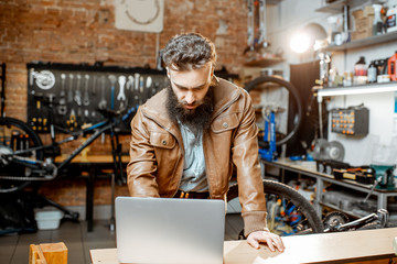 Stylish bearded businessman as a shop owner or manager working with laptop at the bicycle workshop