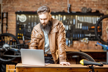Stylish bearded businessman as a shop owner or manager working with laptop at the bicycle workshop