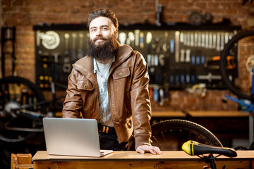 Portrait of a handsome bearded man as bicycle store owner or manager standing with laptop at the bicycle workshop - Powered by Adobe