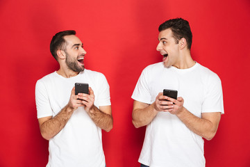 Image of two optimistic men friends 30s in white t-shirts smiling and holding smartphones together