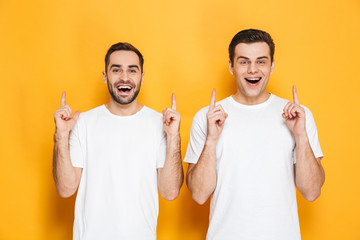 Image of two beautiful men friends 30s in white t-shirts smiling and pointing fingers upward at...
